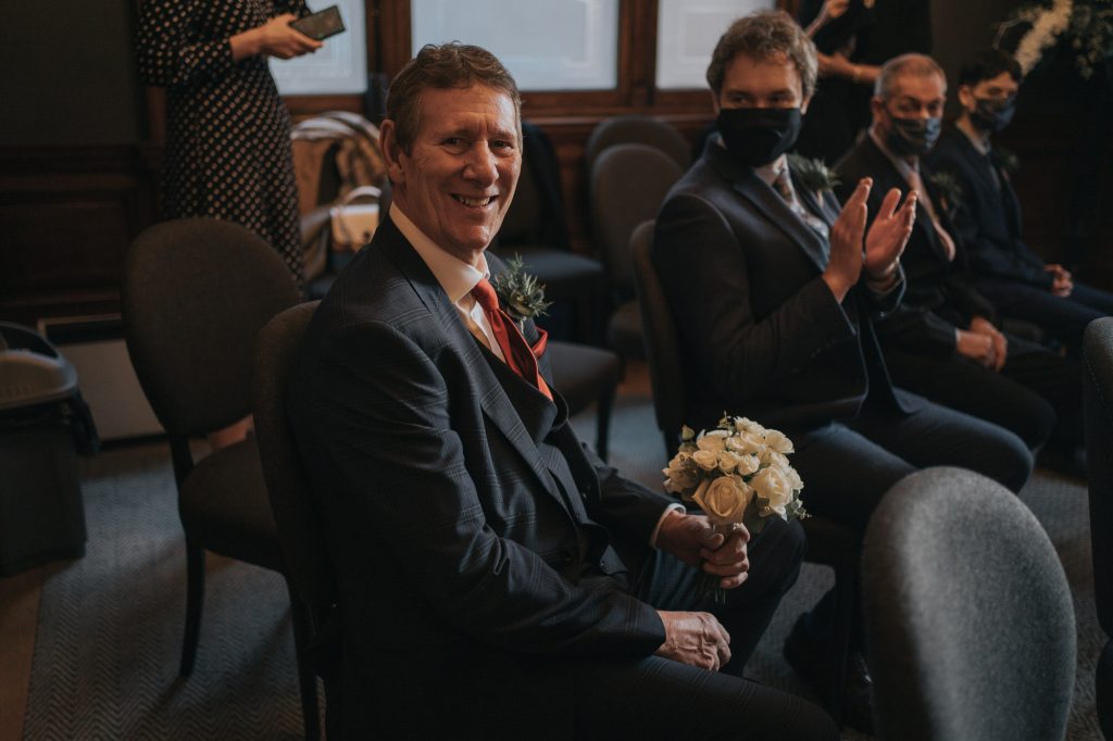The grooms Dad smiles as he holds a bouquet - people are clapping in the background