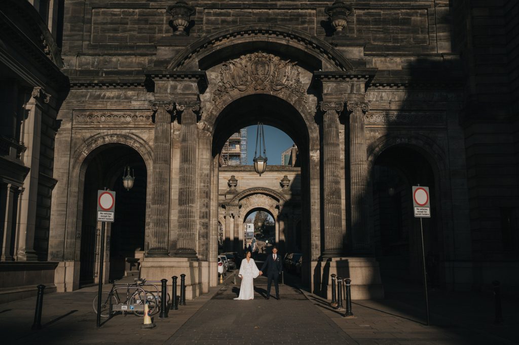 The bride and groom are dwarfed 
 against the immense architecture of Glasgow City Chambers
