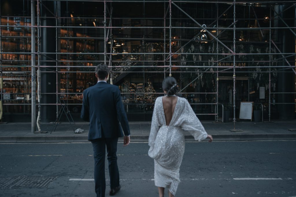 The bride and groom are running across the road towards some Scaffolding