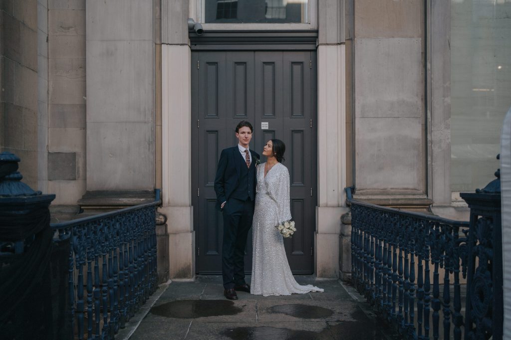The bride and groom pose in front of a grey doorway