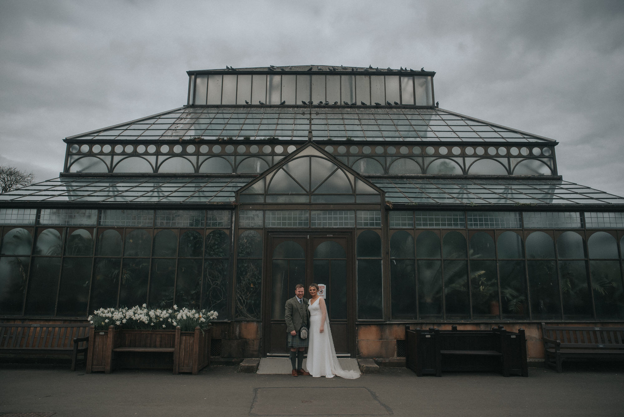 The bride and groom stand in front of a large, ornate green house