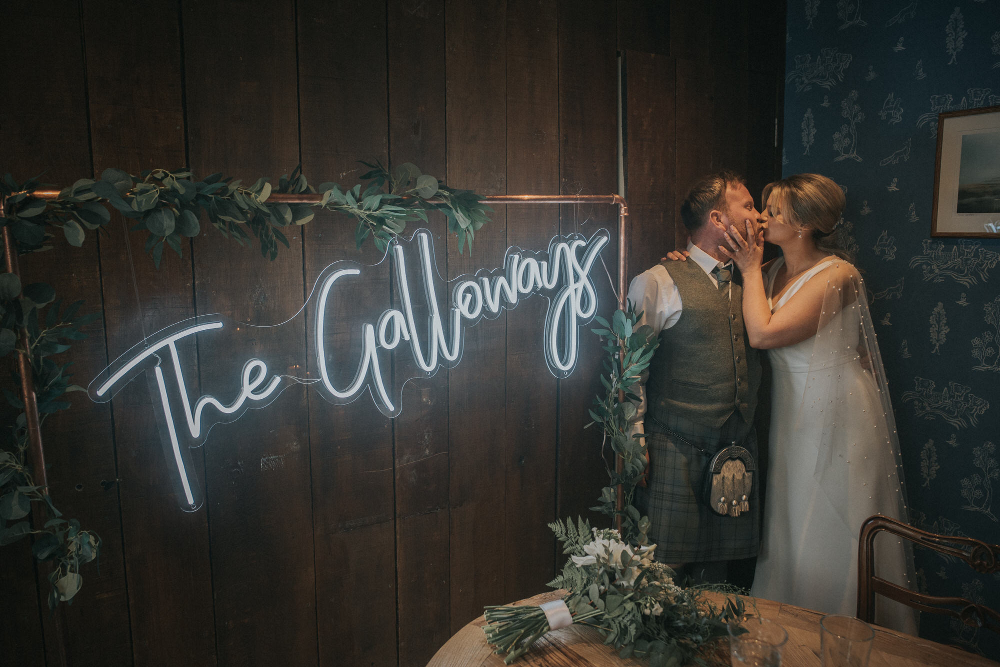 The bride and groom kiss beside the neon The Galloways Sign