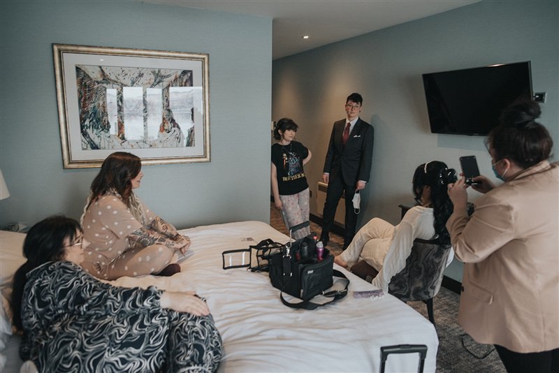 A bridal party sits in a hotel bedroom watching on as the bride gets her hair done - the hairdresser is taking a photo on her phone