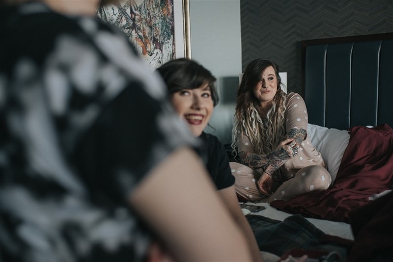 Bridesmaids laugh as they sit on a bed waiting to get their make up done
