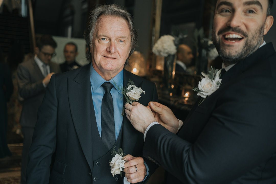 The groom looks behind and laugh as he helps apply the father of the brides buttonhole flower