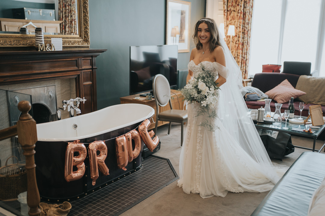 The bride smiles as she stands next to a bath that has balloons on it - the balloons read B R I D E