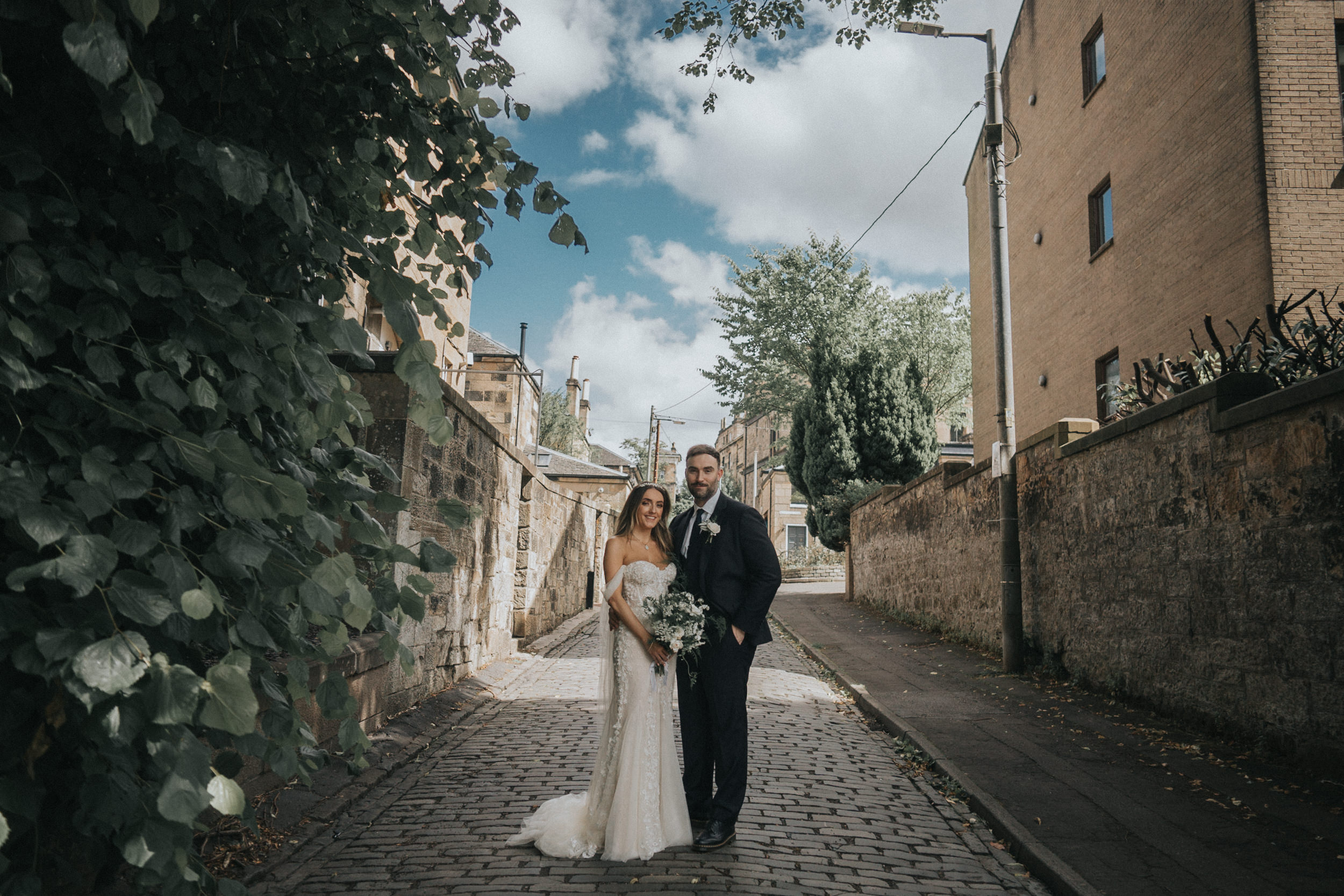 A bride and groom stand in an alleyway behind Hotel Du Vin in Glasgow. The sun is shining, its a beautiful blue sky and the greenery surrounds them beautifully