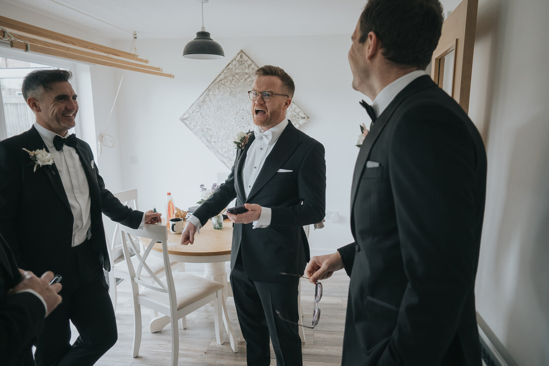 The groom makes a funny angry face as he tells a story while he enjoys his morning with his groomsmen - they are all wearing tuxedos