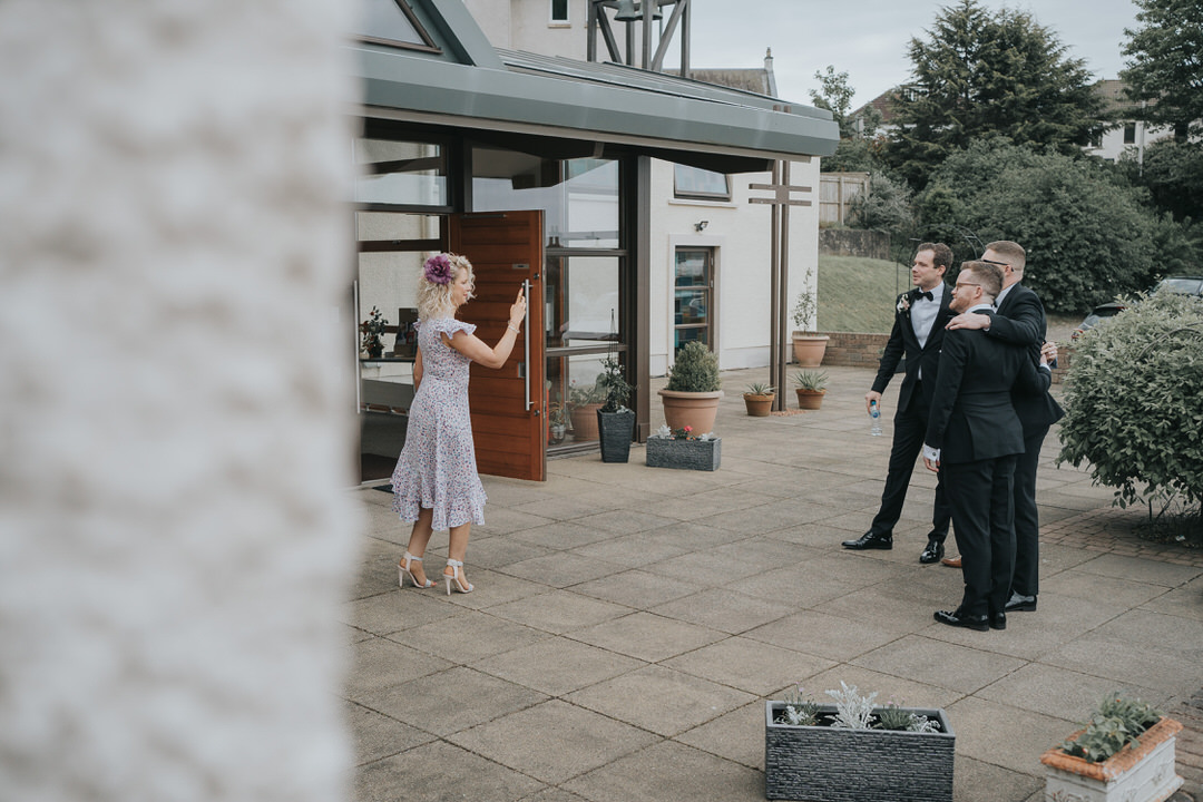 A guest in a purple dress uses her mobile phone to take a photo of the groom and his groomsmen outside the church