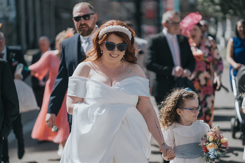The bride is wearing sunglasses and smiles as she walks along in the sunshine whilst holding a small girls hand