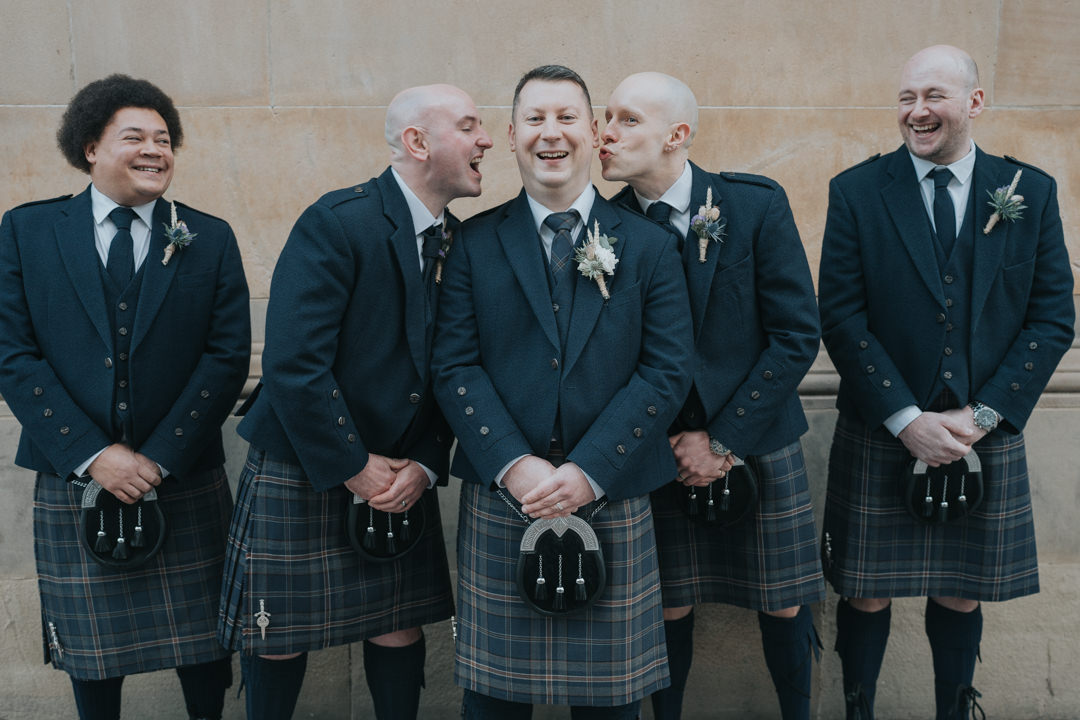 The groom laughs as two of his groomsmen kiss him on the face outside of Citation in Glasgow