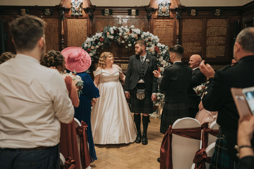 A bride and groom hold hands as they walk down the aisle after their Trades Hall Glasgow Wedding